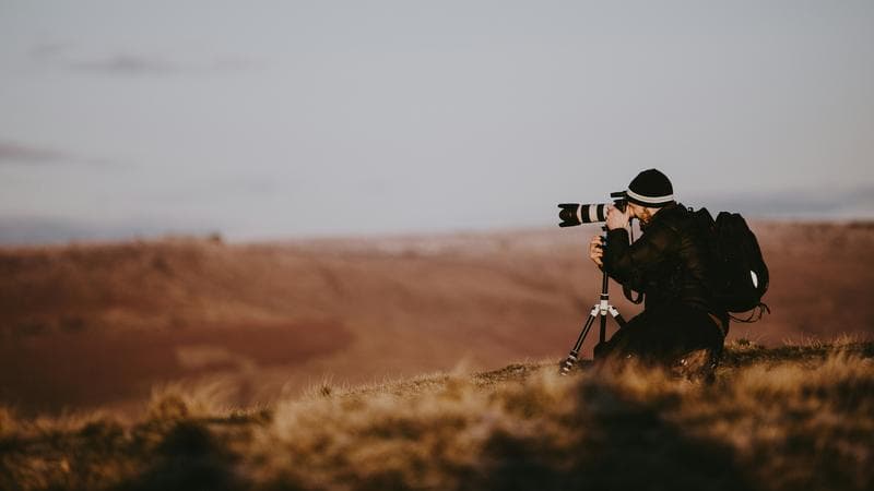 A photographer kneeling behind a tripod on a moor at dusk