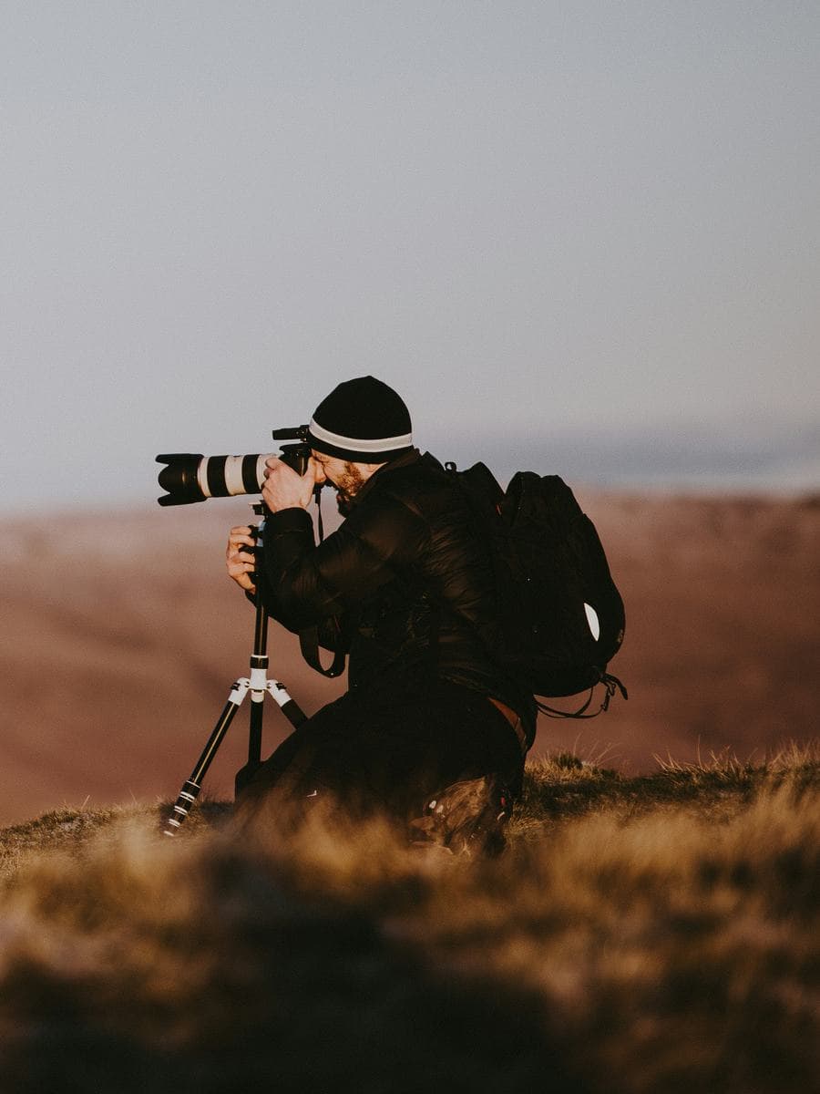 A photographer kneeling behind a tripod on a moor at dusk, looking out over the hills
