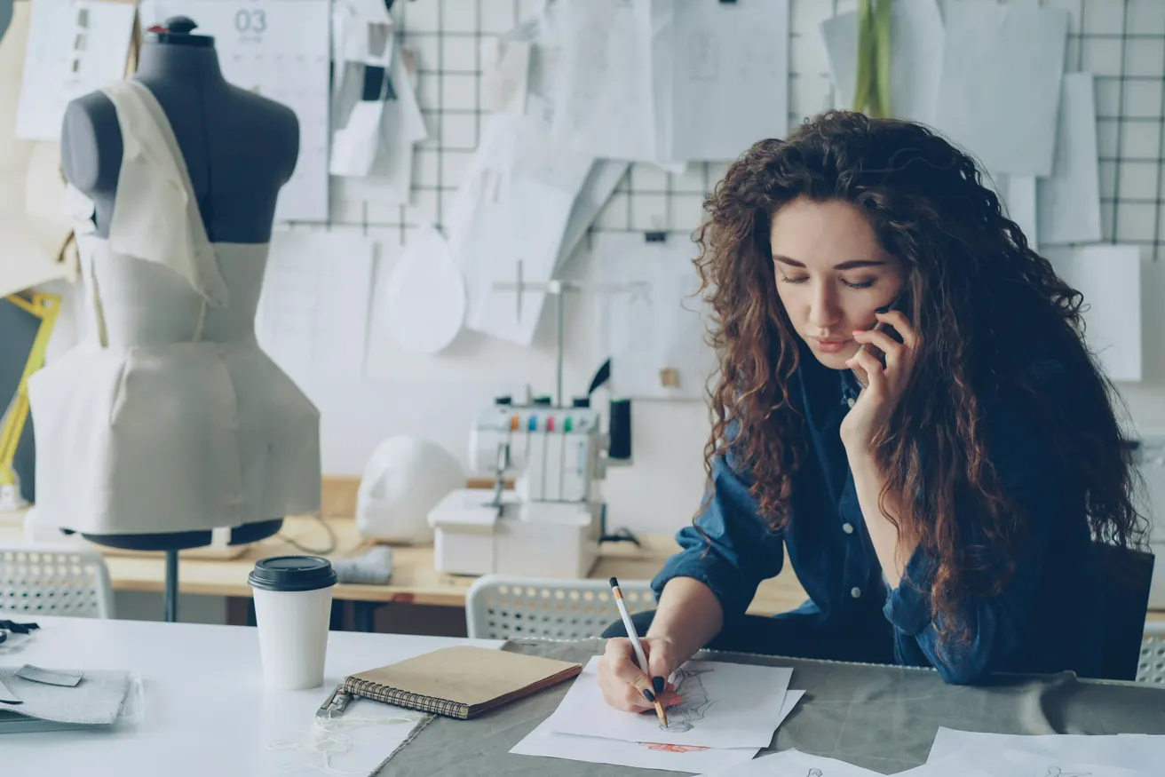 A dressmaker on the phone, sketching at her worktable beside a dress form